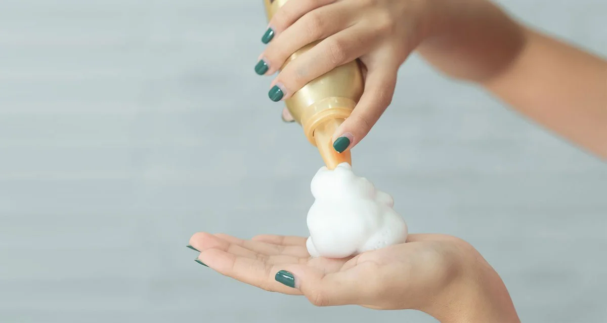 Close-up of hands with teal nails dispensing foam soap onto a palm from a bottle.